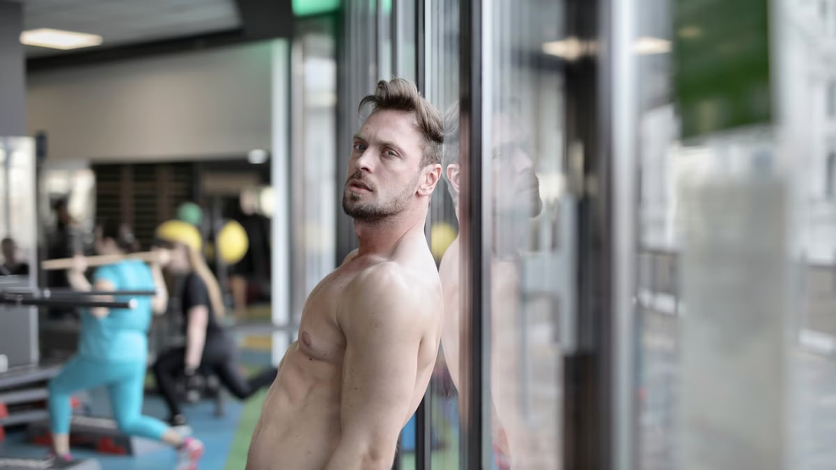 Shirtless man reflects at gym window, showcasing muscular physique and determination.
